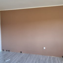 Usługi Remontowe Marcin Frączak - Pokój po częściowym odświeżeniu: jedna ściana pomalowana na brązowo, druga w odcieniach bieli, nowe panele podłogowe z czarnymi klinami dystansowymi przy ścianie.