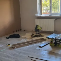 Układanie paneli podłogowych w pokoju, widoczne narzędzia: piła ukośnica, piła stołowa, miarka, rozłożone panele Kronostep, brązowa ściana w tle, dwa okna w górnej części kadru.