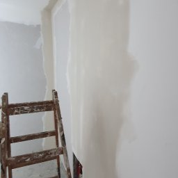 Usługi Remontowe Marcin Frączak - Ściana w trakcie prac wykończeniowych, widoczne świeżo nałożone warstwy, drewniana drabina oparta o ścianę, podłoga z resztkami materiałów budowlanych i śrubokrętem.