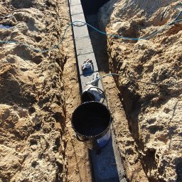 Usługi Remontowe Marcin Frączak - Przygotowanie fundamentów: Wąski wykop z betonowymi bloczkami, zabezpieczonymi czarną masą bitumiczną w wiadrze, widoczne rękawice robocze i fragment ubrania roboczego