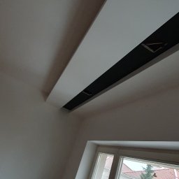 Usługi Remontowe Marcin Frączak - Biały sufit z czarną wnęką oświetleniową i widokiem na okno z dwoma brązowymi torbami i czarnym wentylatorem stojącym na parapecie.
