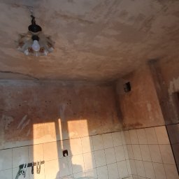 Usługi Remontowe Marcin Frączak - Remontowana kuchnia: Widok na surowy sufit z widocznymi śladami prac, fragment ściany z resztkami tynku i starej glazury, blat kuchenny z pozostałościami materiałów budowlanych i narzędziami.