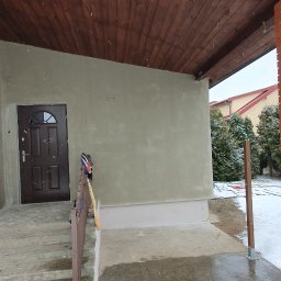 Usługi Remontowe Marcin Frączak - Świeżo otynkowana elewacja domu jednorodzinnego z drewnianym podbitkiem, widoczne narzędzia i niedokładnie posprzątany teren wokół budynku w zimowej scenerii.