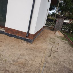 Usługi Remontowe Marcin Frączak - Świeżo otynkowana ściana domu z zamontowanym systemem odprowadzania wody deszczowej, widoczne przygotowania do prac ziemnych, wytyczone sznurki i wbite słupki.