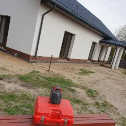 Świeżo otynkowany dom jednorodzinny z ciemną dachówką, widoczna czerwona skrzynka narzędziowa i niwelator laserowy na ławce, teren wokół domu w trakcie zagospodarowywania.