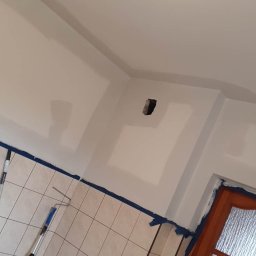 Usługi Remontowe Marcin Frączak - Świeżo zagruntowana ściana w łazience z widocznym otworem na gniazdko elektryczne, zabezpieczonymi listwami przypodłogowymi i fragmentem drewnianych drzwi z szybą ornamentową.