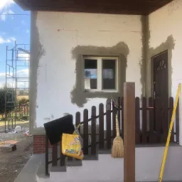 Elewacja domu w trakcie prac ociepleniowych, widoczne płyty styropianowe, zaprawa wokół okna, drabina rusztowania w tle, drewniane wykończenie podbitki dachowej, bosa para butów zwisa z ogrodzenia.