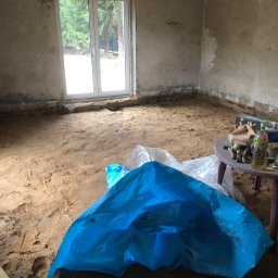 Usługi Remontowe Marcin Frączak - Przygotowanie do remontu pokoju: zerwana podłoga, widoczne ślady wilgoci na ścianach, narzędzia i materiały budowlane na okrągłym stoliku, zabezpieczająca folia w kolorze niebieskim na podłodze.