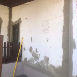 Usługi Remontowe Marcin Frączak - Ściana budynku w trakcie ocieplania styropianem, widoczne płyty styropianowe, częściowo pokryte szarą warstwą kleju, żółta poziomica oparta o ścianę, fragment drewnianego płotu i drzwi wejściowe...