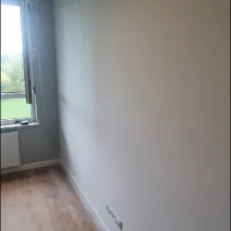 Pomalowane na jasno ściany pokoju z drewnopodobną podłogą, widoczne gniazdka elektryczne w dolnej części ściany i okno z widokiem na zieleń za nim.