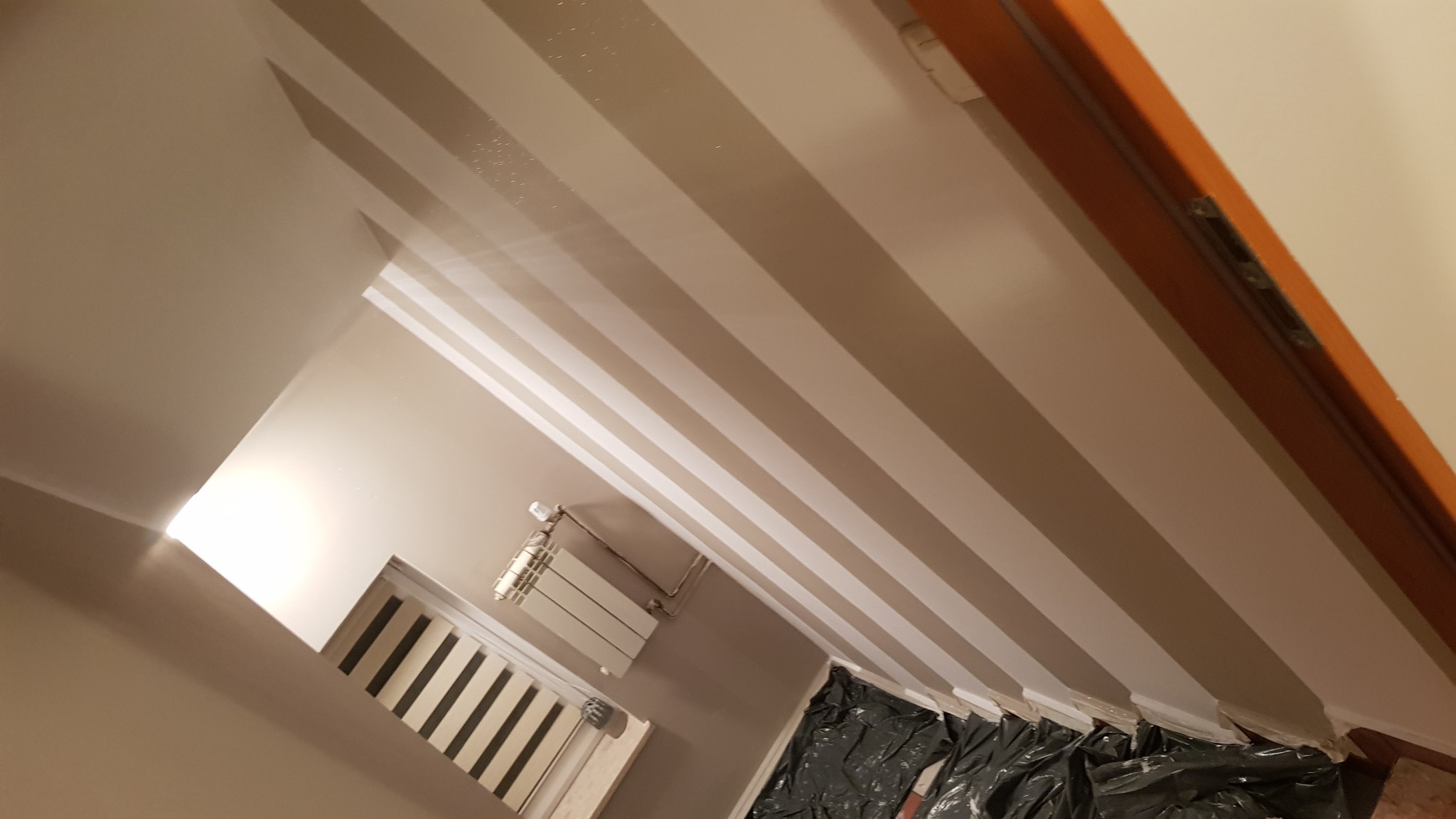 Świeżo pomalowany pokój z beżowymi ścianami i białymi pasami, grzejnik i okno widoczne w tle, zabezpieczona folią podłoga.