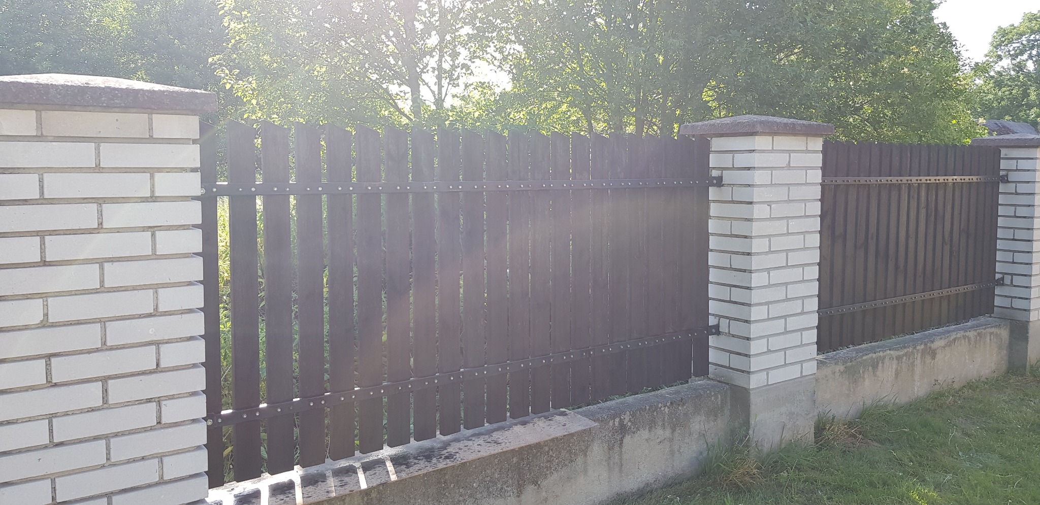 Nowoczesne ogrodzenie z ciemnego drewna z elementami metalowymi i białymi słupkami z cegły, widoczne zieleń w tle.