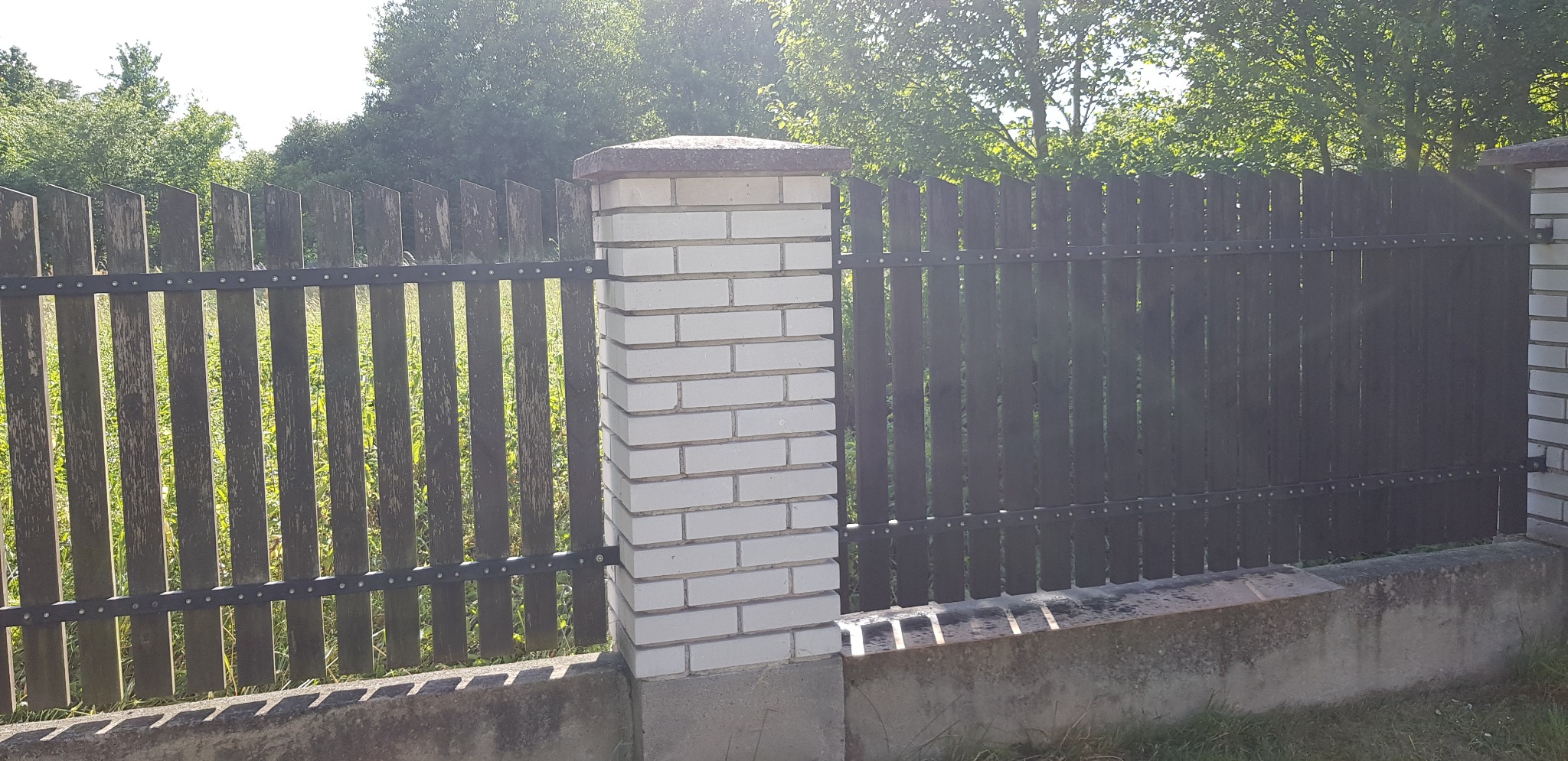 Drewniany płot z pionowymi deskami, mocowany na metalowych szynach, wsparty na betonowej podmurówce i zakończony białym, murowanym słupkiem z betonową czapką.