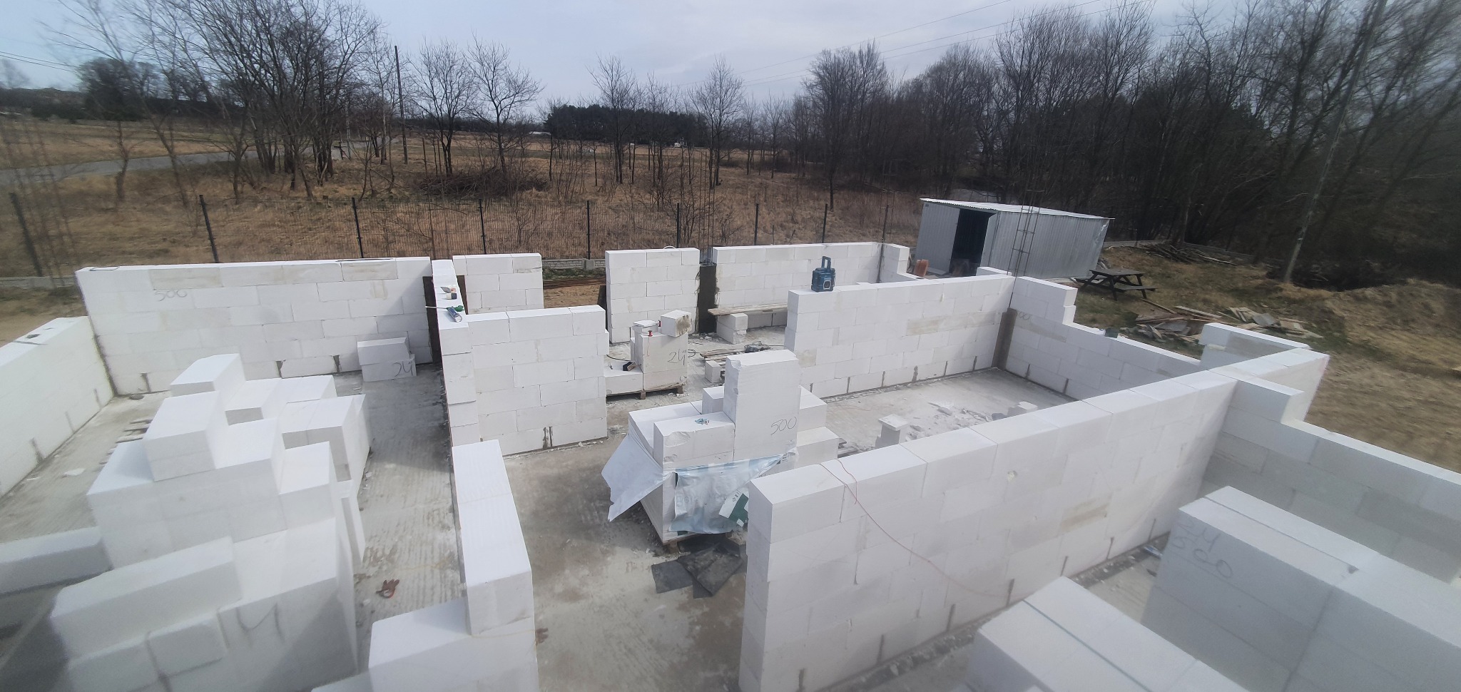 Wznoszenie ścian parteru domu z białych bloczków, widok z góry na plac budowy z zewnątrz, w tle drzewa i blaszany garaż.