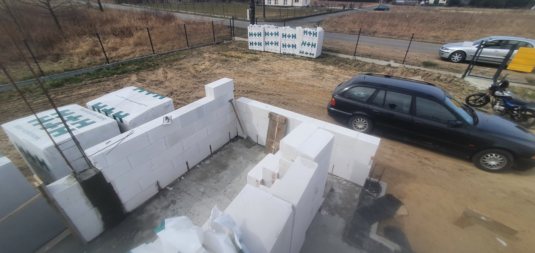 Wznoszenie ścian parteru z bloczków betonu komórkowego na placu budowy, widoczne palety z zapasem materiału H+H, czarny samochód kombi i motocykl w tle.