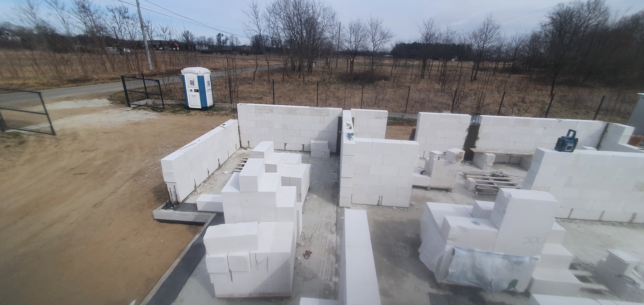 Wznoszenie ścian parteru domu z białych bloczków, widoczna przenośna toaleta na zewnątrz, teren budowy w pochmurny dzień.