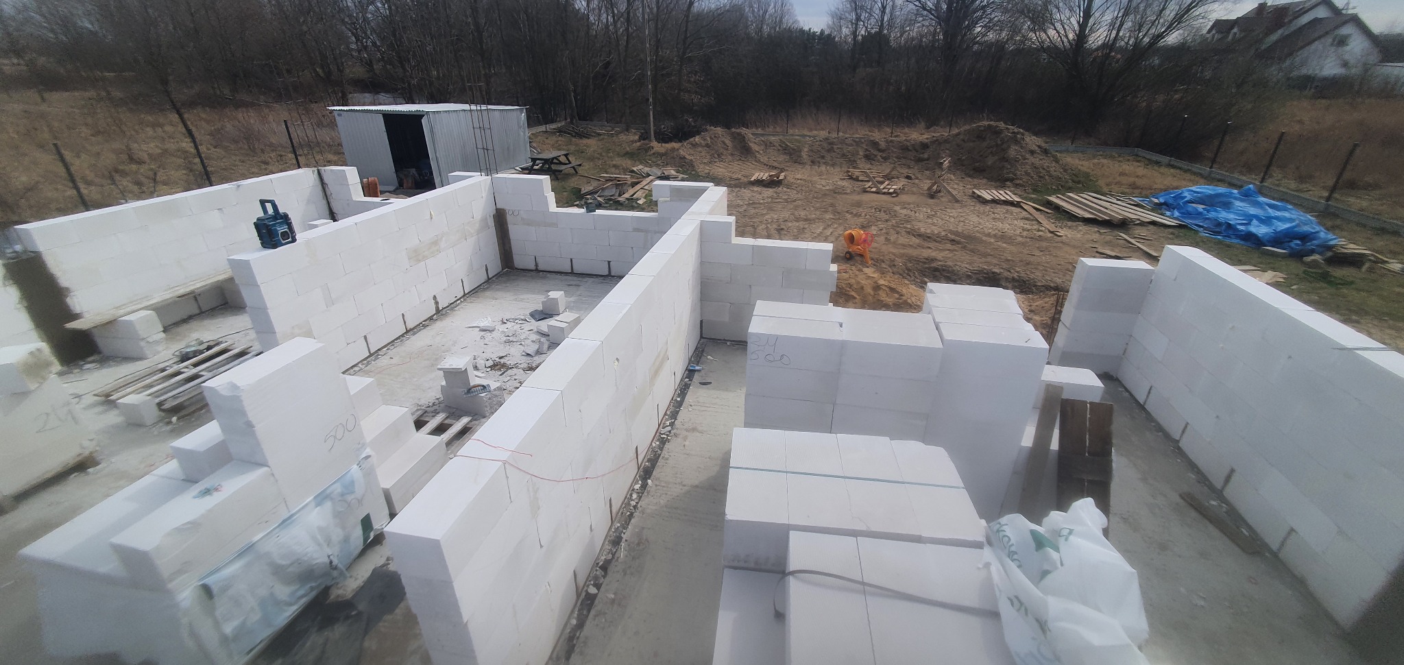 Budowa domu jednorodzinnego w trakcie wznoszenia ścian z białych bloczków betonu komórkowego, widok z góry na fundamenty i pierwsze warstwy muru, z narzędziami budowlanymi i stertą materiałów w tle.