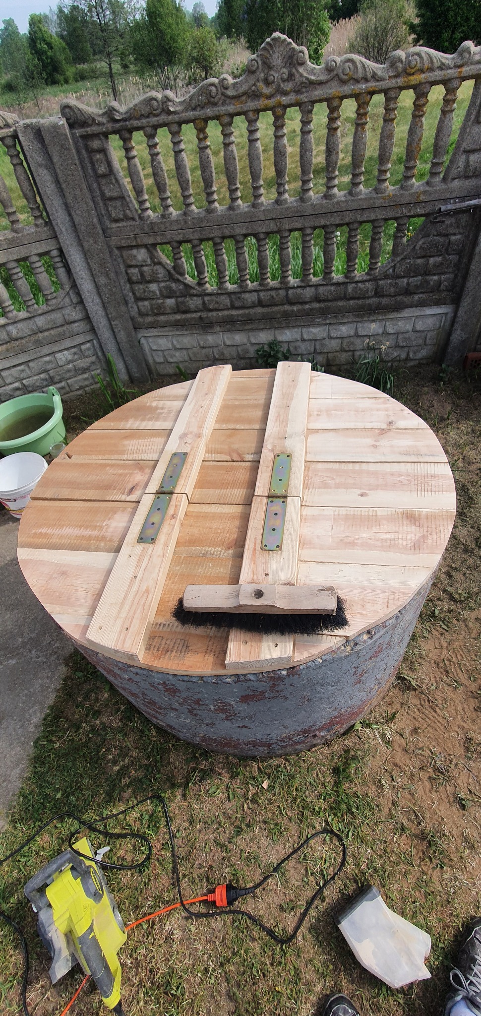Okrągła studnia z drewnianą pokrywą, zabezpieczoną metalowymi zawiasami, obok leży szczotka do zamiatania, a w tle widać betonowe ogrodzenie.