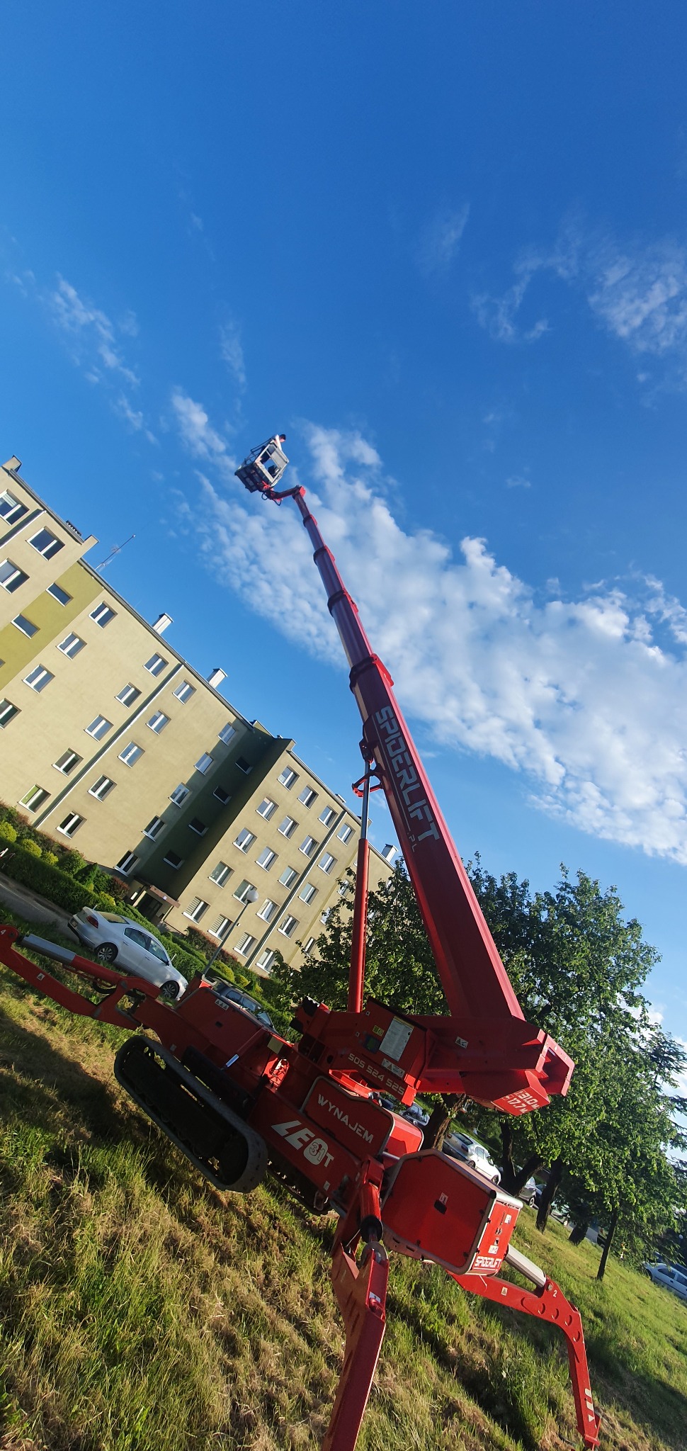 Czerwony podnośnik koszowy Spiderlift na gąsienicach, z wysięgnikiem skierowanym ku górze, na tle bloku mieszkalnego i błękitnego nieba z chmurami.