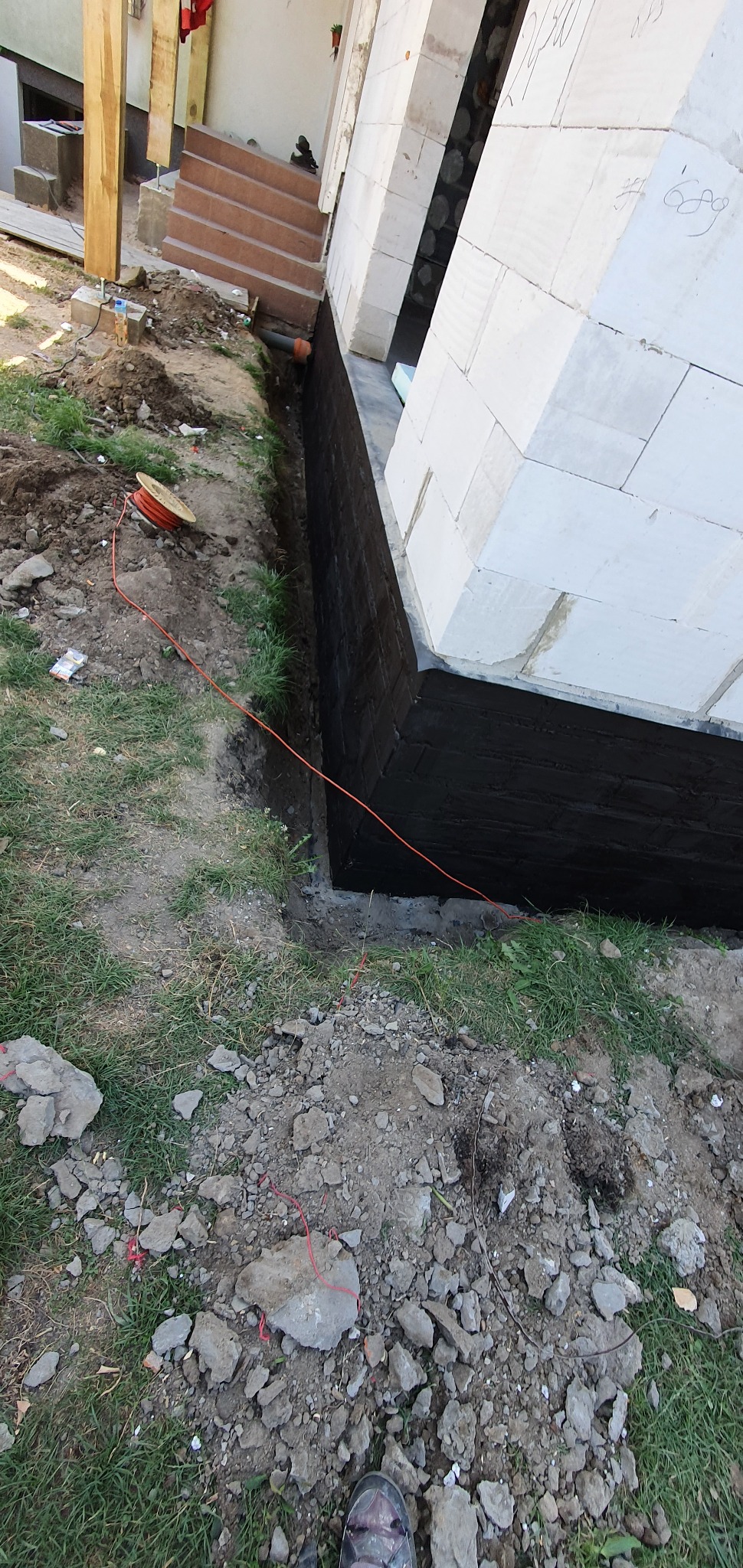 Hydroizolacja fundamentów domu z bloczków betonowych, widoczna czarna powłoka izolacyjna, obok wykop z pomarańczowym przewodem i elementami instalacji.