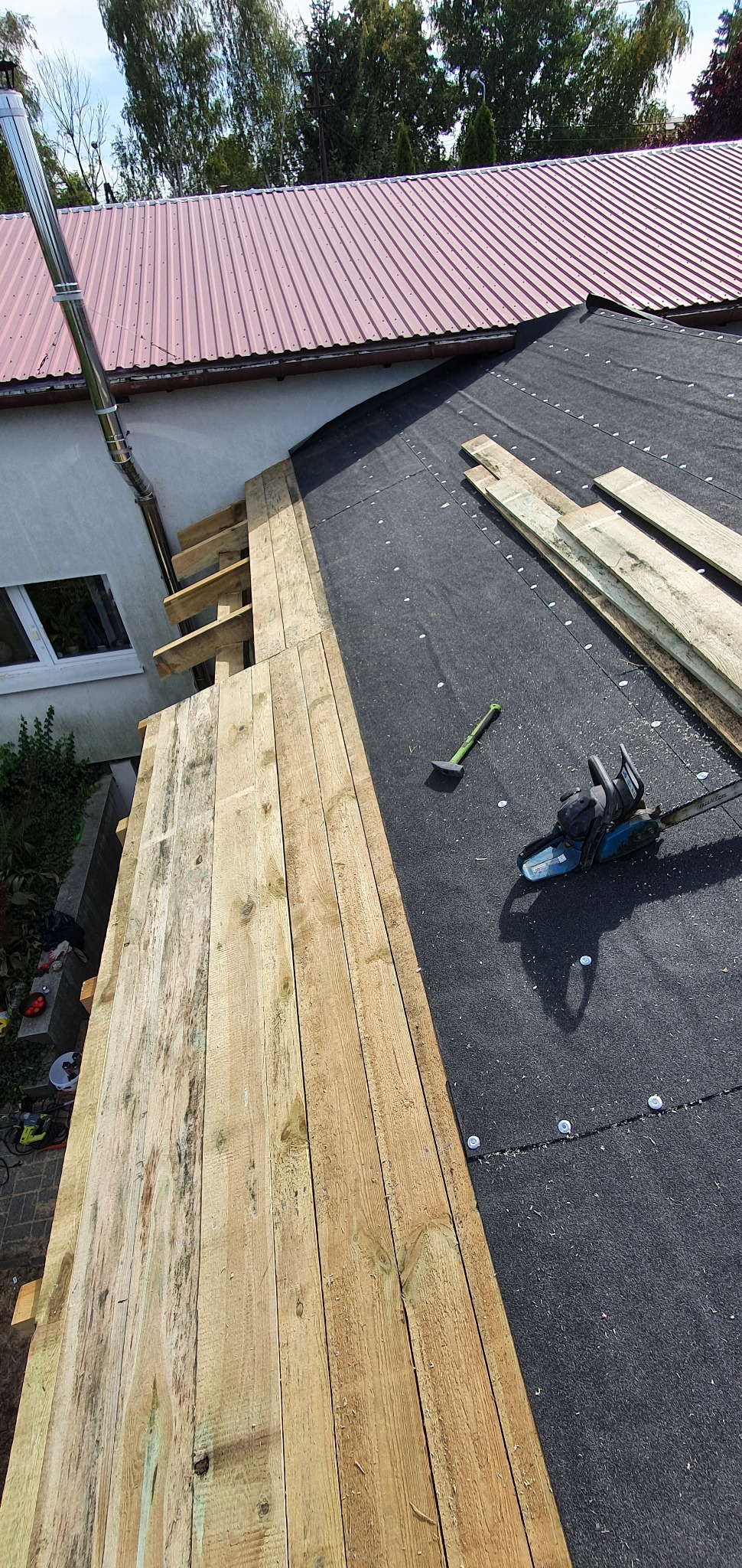 Montaż izolacji na dachu z widoczną membraną, drewnianymi łatami i narzędziami: piła łańcuchowa i młotek dekarski.