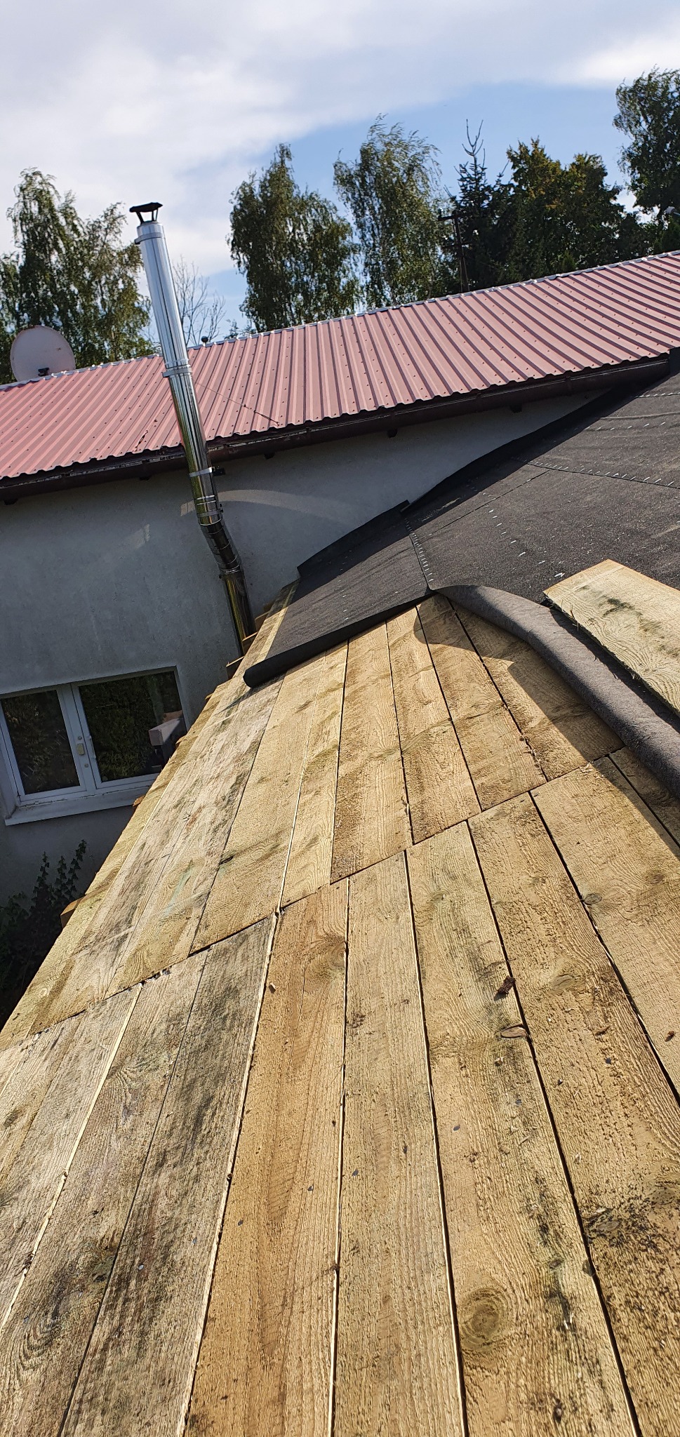 Deskowanie dachu z widoczną membraną i kominem stalowym na tle domu z blachodachówką.