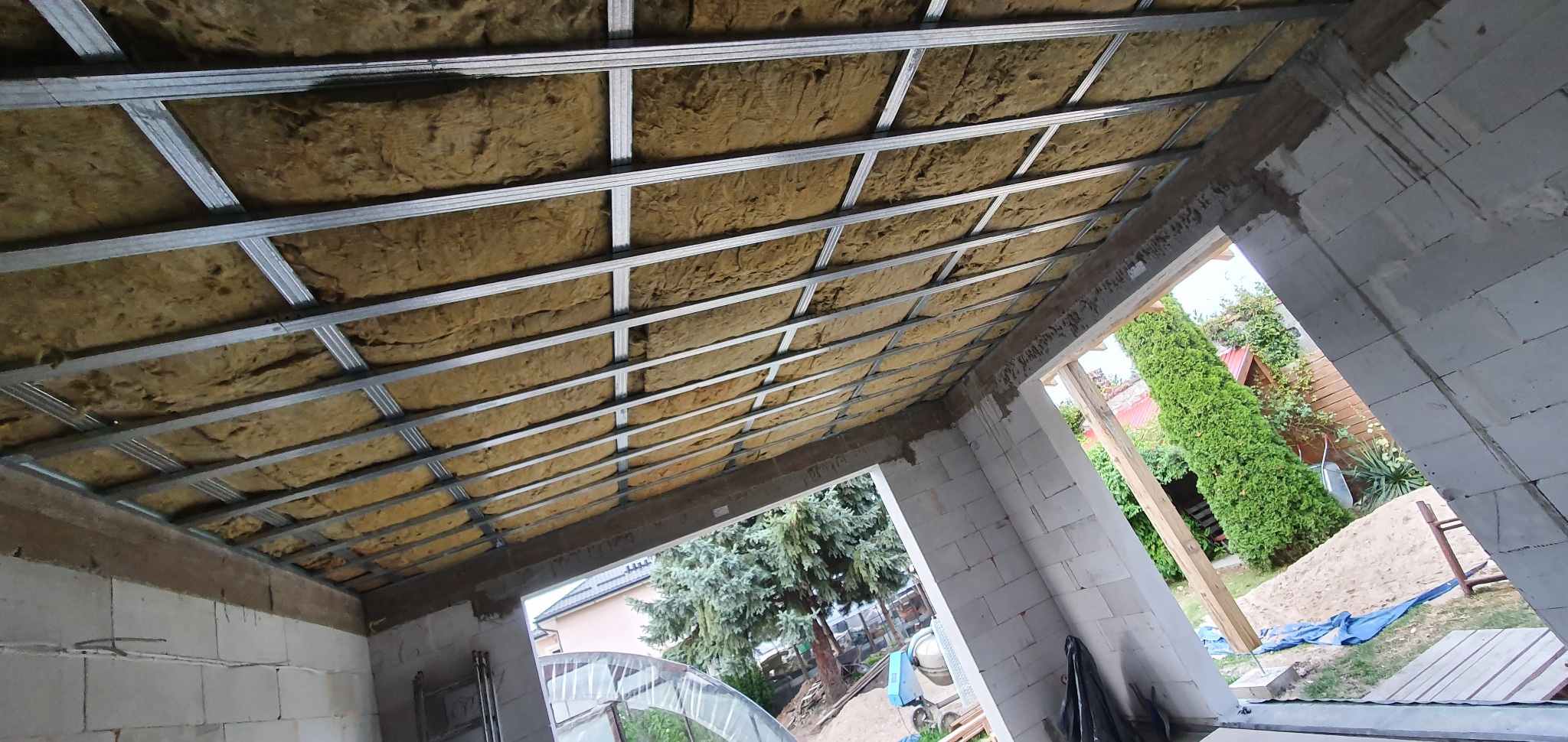 Poddasze w trakcie ocieplania, widoczne metalowe profile i wełna mineralna, ściany z białego bloczka, widok na zewnątrz na drzewa i ogród.