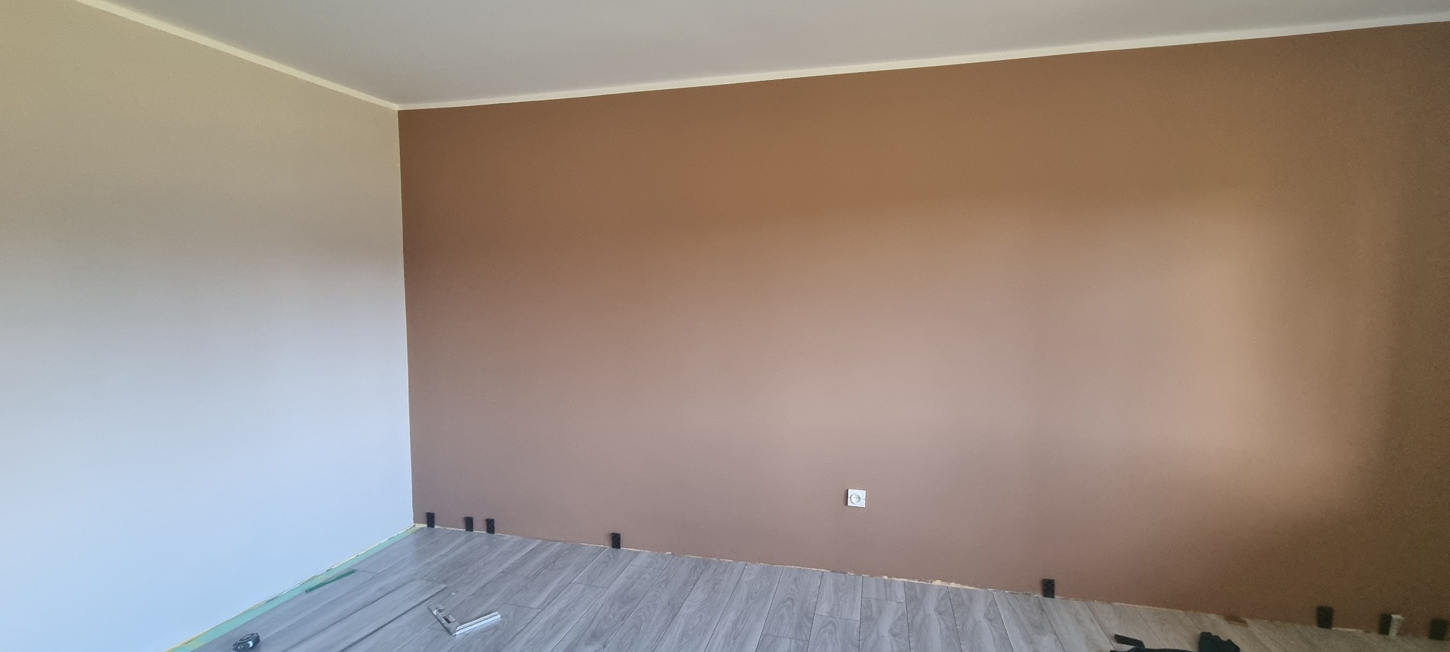 Pokój po częściowym odświeżeniu: jedna ściana pomalowana na brązowo, druga w odcieniach bieli, nowe panele podłogowe z czarnymi klinami dystansowymi przy ścianie.