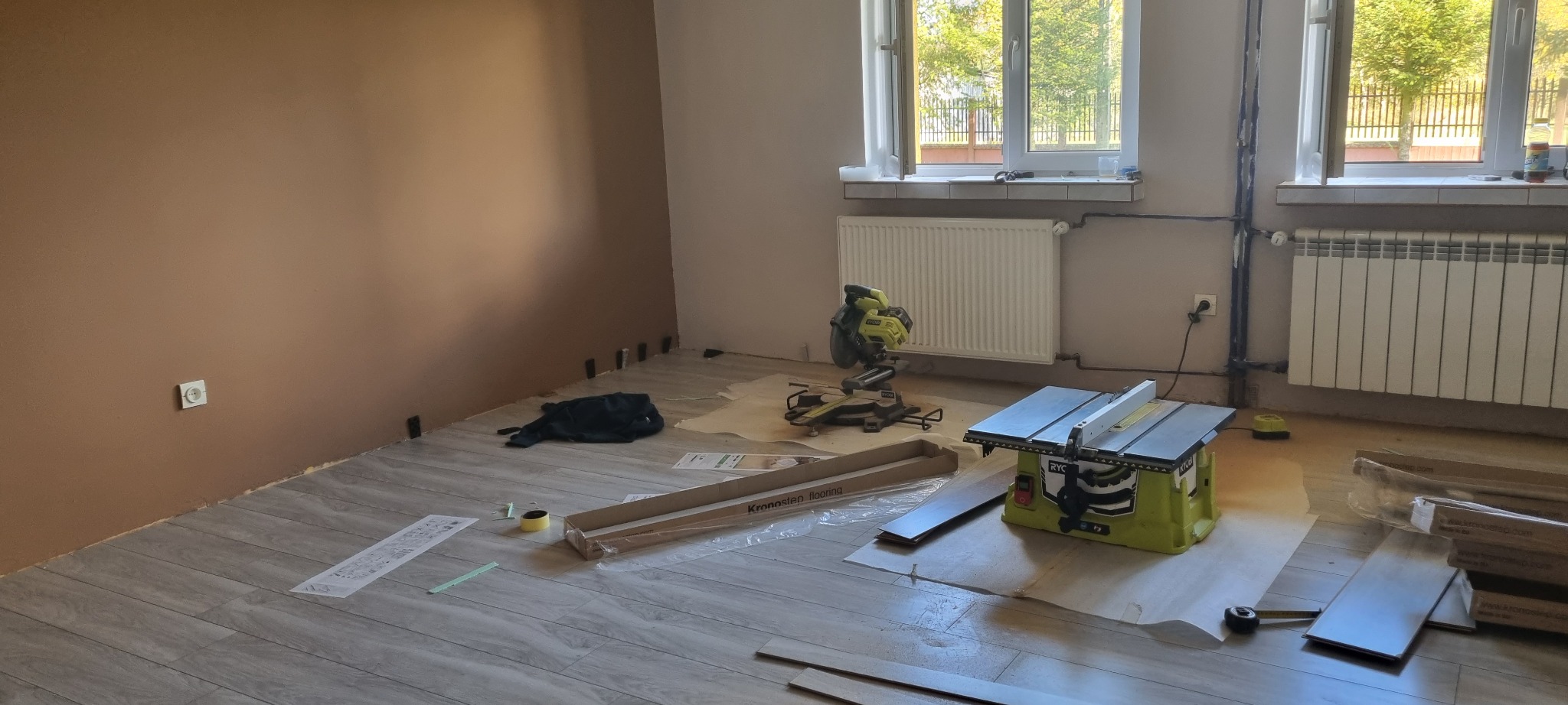 Układanie paneli podłogowych w pokoju, widoczne narzędzia: piła ukośnica, piła stołowa, miarka, rozłożone panele Kronostep, brązowa ściana w tle, dwa okna w górnej części kadru.