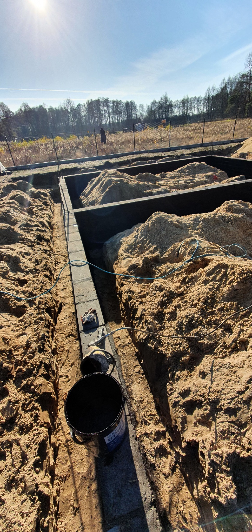 Przygotowanie fundamentów: wykopane rowy, zalane betonem, wypełnione piaskiem. Widoczne wiadro z czarną substancją izolacyjną i rękawice robocze.