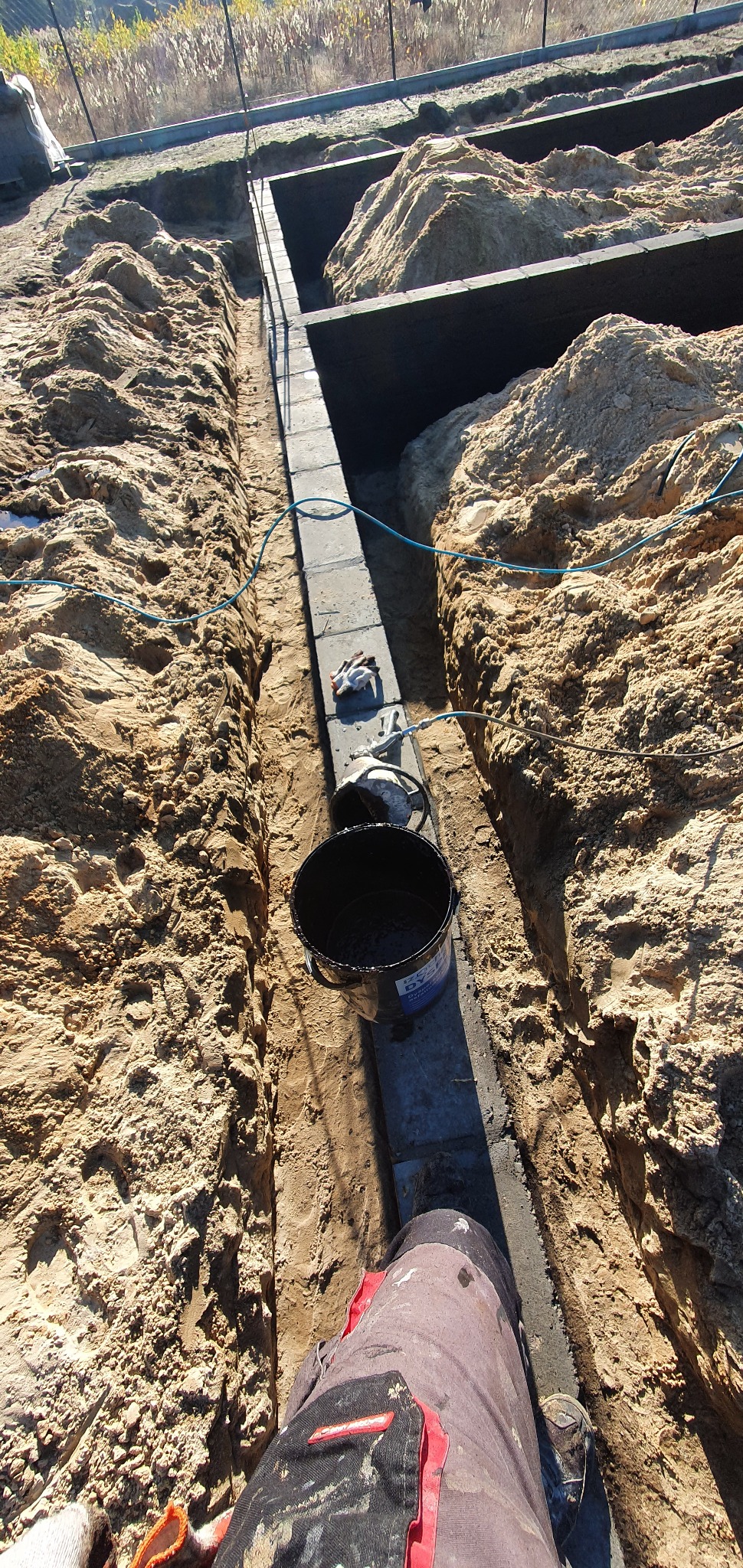 Przygotowanie fundamentów: Wąski wykop z betonowymi bloczkami, zabezpieczonymi czarną masą bitumiczną w wiadrze, widoczne rękawice robocze i fragment ubrania roboczego