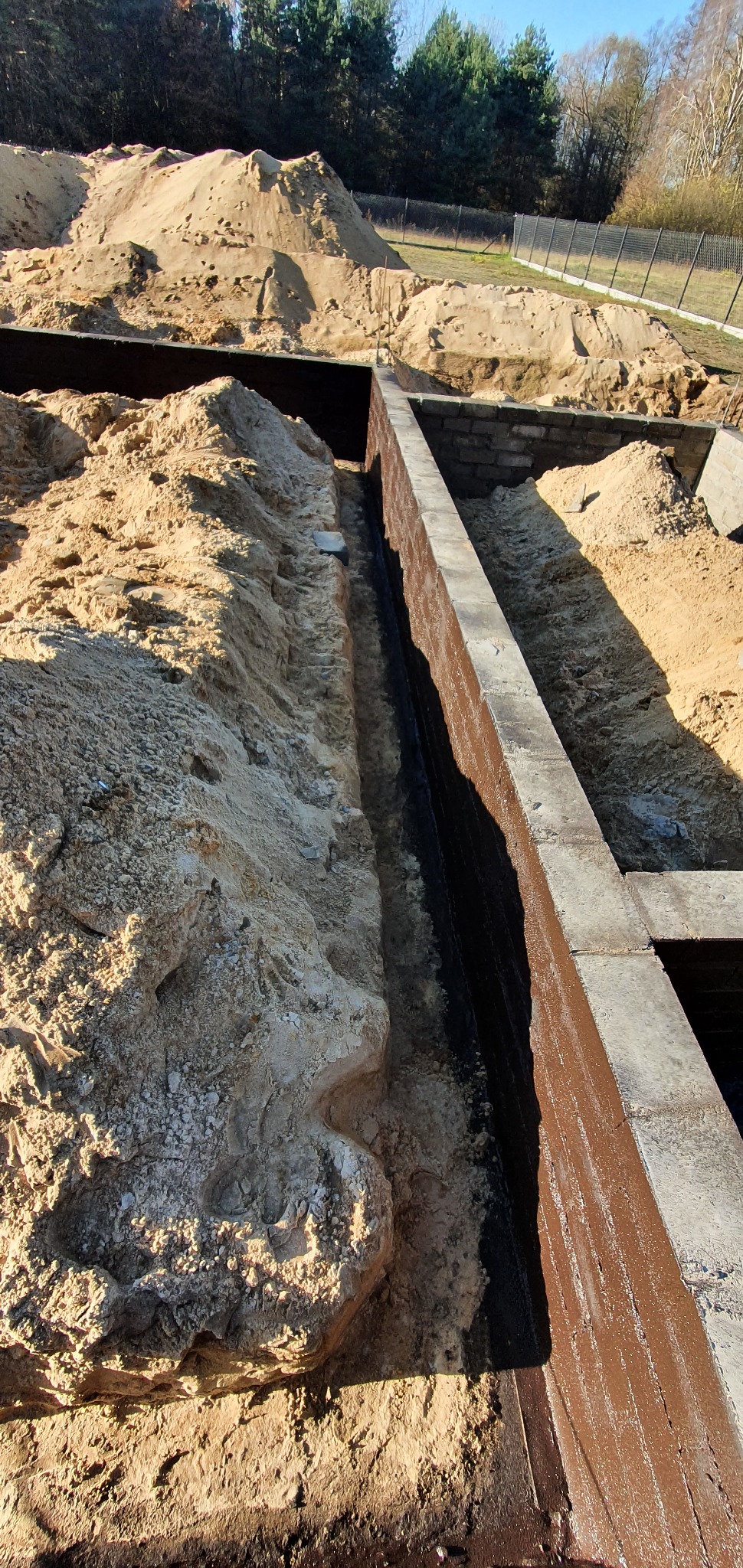 Fundamenty budynku w trakcie budowy, zabezpieczone hydroizolacją, widoczny wykop i nasypana ziemia.