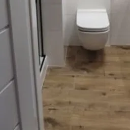 Widok na wykończoną łazienkę z drewnopodobnymi płytkami podłogowymi, białą wiszącą toaletą i fragmentem kabiny prysznicowej z białymi płytkami na ścianach, uchwycony z perspektywy wejścia.