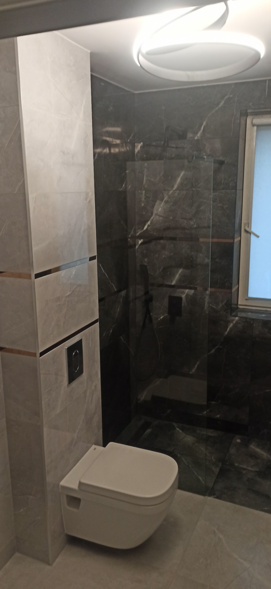 Nowoczesna łazienka z wiszącą miską WC, szklaną kabiną prysznicową i marmurowymi płytkami w odcieniach szarości, oświetlona okrągłą lampą sufitową.