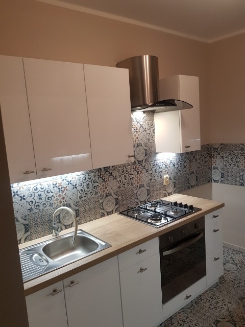 Nowoczesna kuchnia z białymi szafkami, drewnianym blatem i dekoracyjnymi płytkami nad zlewem i kuchenką gazową, oświetlenie LED pod szafkami.