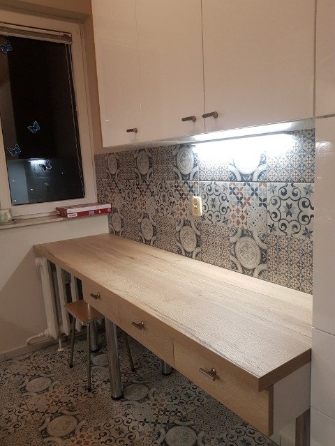 Jasna drewniana deska z szufladami wsparta na metalowych nogach, dopasowana do wzorzystych płytek na ścianie i podłodze, białe szafki wiszące nad blatem, widoczne dekoracje okienne.