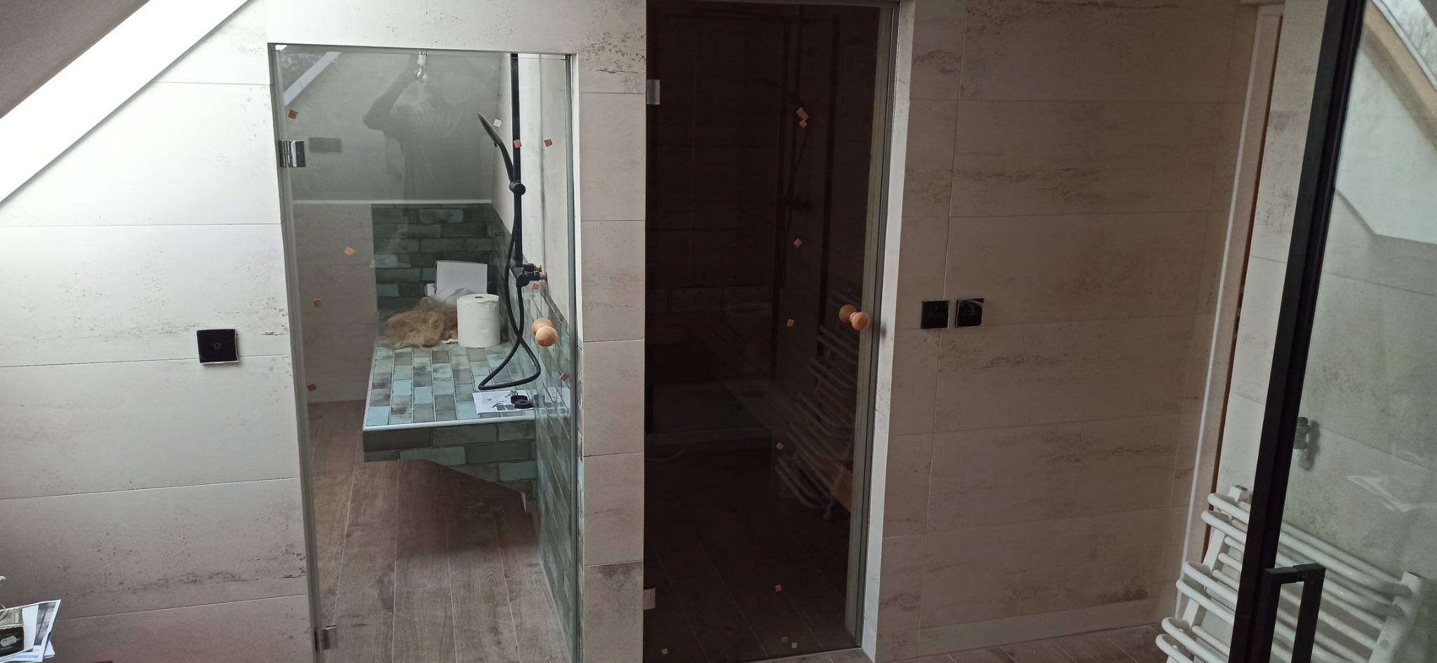 Łazienka na poddaszu w trakcie remontu: widoczna kabina prysznicowa ze szklanymi drzwiami, fragment sauny, płytki imitujące drewno na ścianach, niebiesko-szara mozaika na wnęce, czarna armatura...