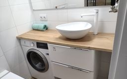 Nowoczesna łazienka z umywalką nablatową w kształcie misy, białą baterią, blatem z jasnego drewna nad pralką i szafką, białe płytki na ścianie, zielony ręcznik.