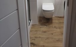 Widok na wykończoną łazienkę z drewnopodobnymi płytkami podłogowymi, białą wiszącą toaletą i fragmentem kabiny prysznicowej z białymi płytkami na ścianach, uchwycony z perspektywy wejścia.