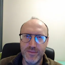 Portret mężczyzny w okularach, siedzącego na czarnym krześle na tle jasnej ściany, z widocznymi refleksami na szkłach.