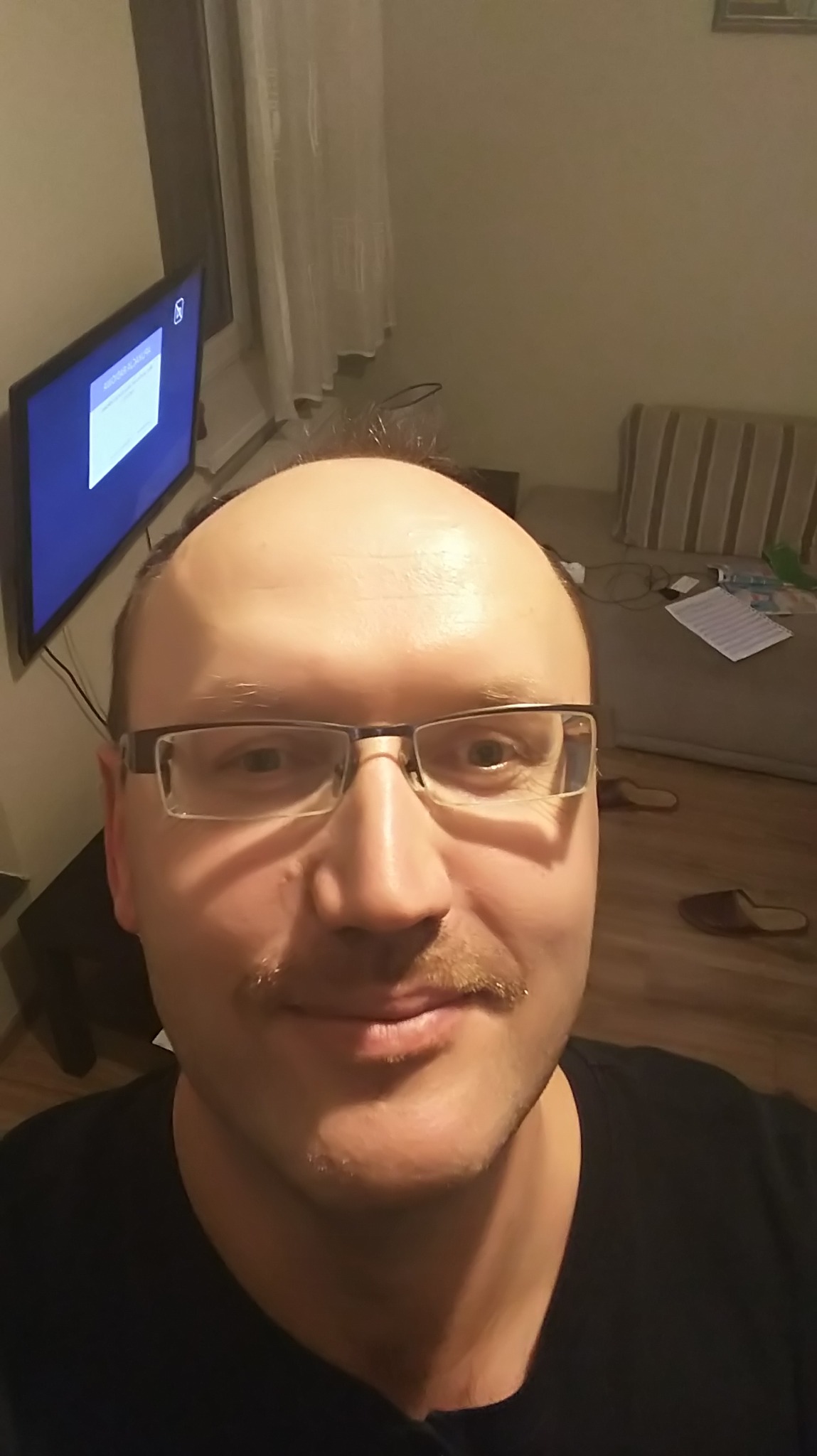 Portret mężczyzny w okularach z wąsami, uśmiechniętego, z telewizorem i kanapą w tle. Nietypowy kadr z bliska.