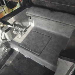 Wnętrze kabiny pojazdu rolniczego lub specjalistycznego z szarą skórą ekologiczną, widoczne elementy sterowania i podłoga z teksturą.