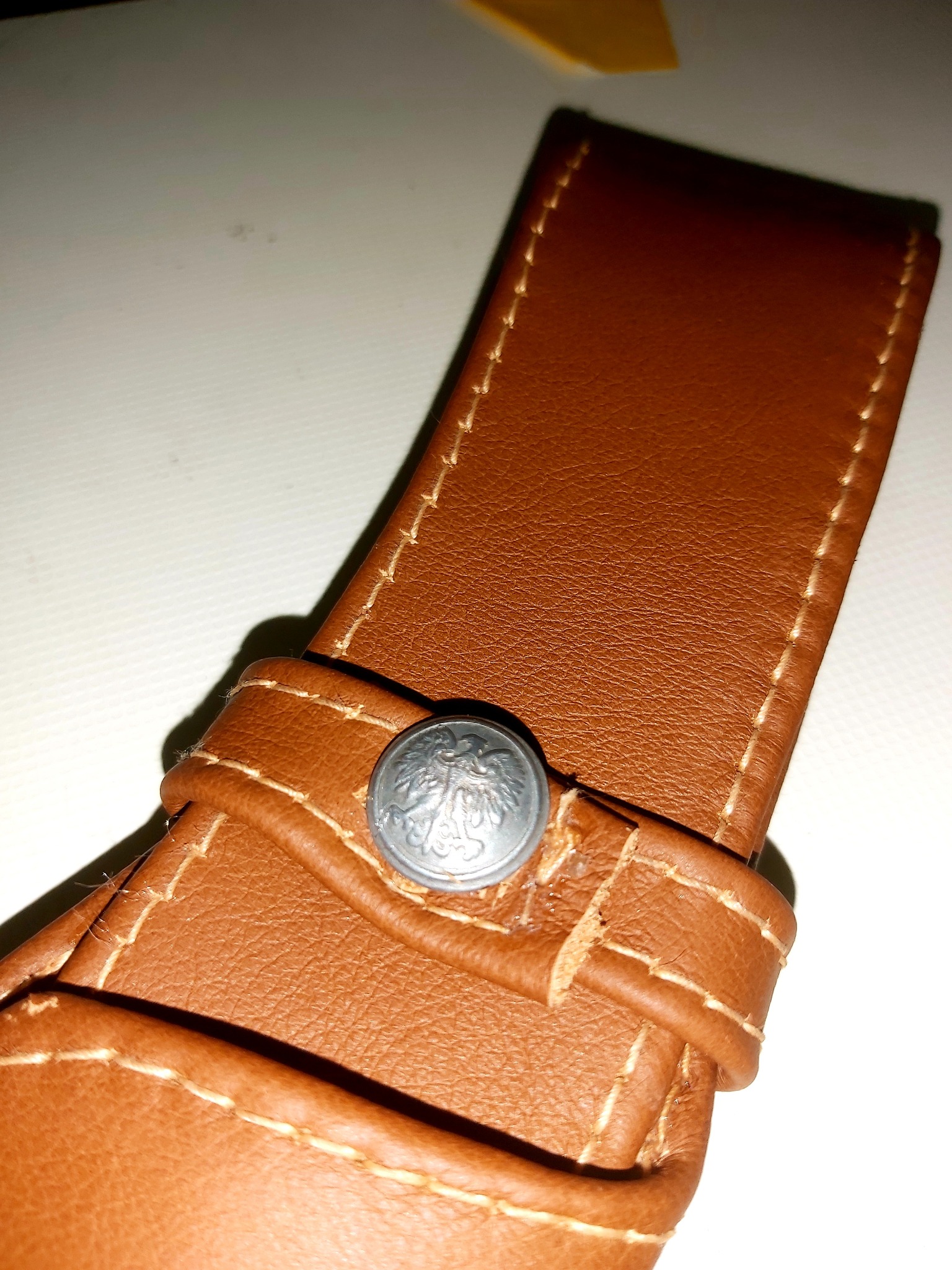 Brązowy skórzany pasek z ozdobnym srebrnym guzikiem z motywem orła, widoczne precyzyjne przeszycia jasną nicią.