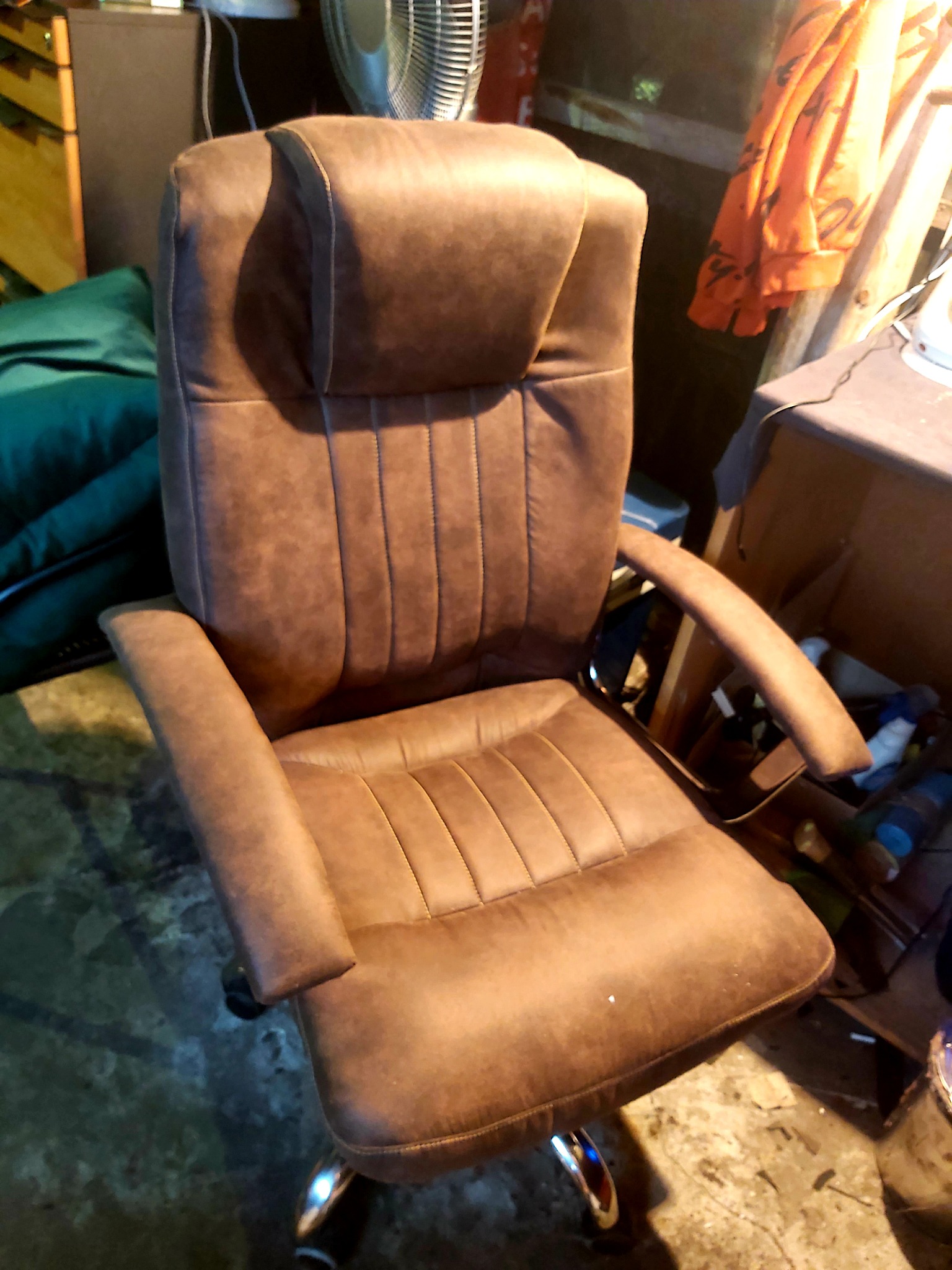 Brązowe krzesło biurowe z wysokim oparciem i zagłówkiem, tapicerowane materiałem przypominającym zamsz, na kółkach, sfotografowane w pomieszczeniu z widocznym bałaganem.