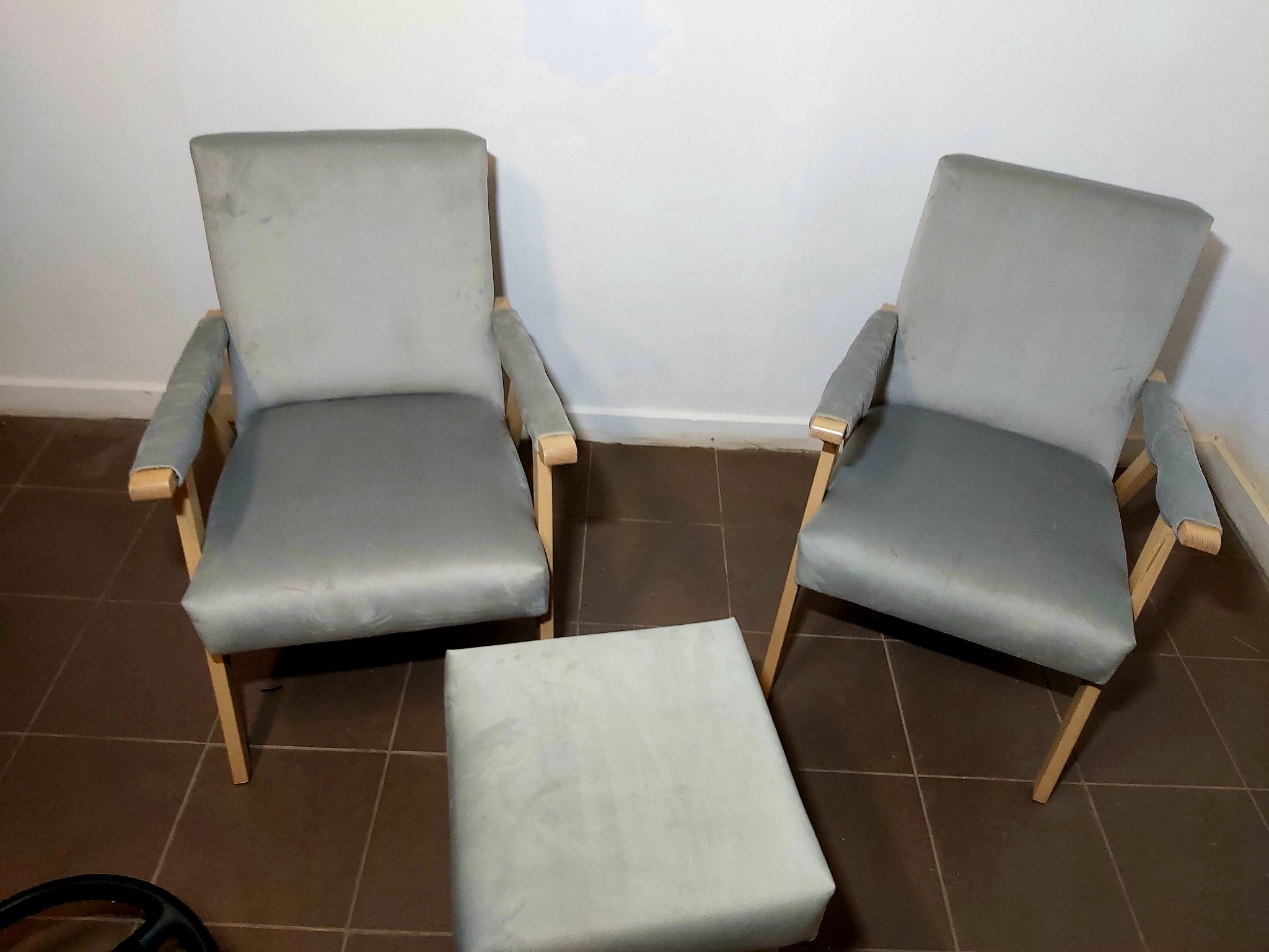 Dwa szare, tapicerowane fotele klubowe z drewnianymi nóżkami i podnóżek na brązowej podłodze z płytek.