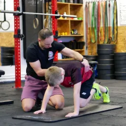 Mężczyzna w czarnej koszulce z logo asekuruje chłopca podczas ćwiczenia na czarnej macie w siłowni; w tle widoczne sprzęty do ćwiczeń, w tym sztanga i drabinki.