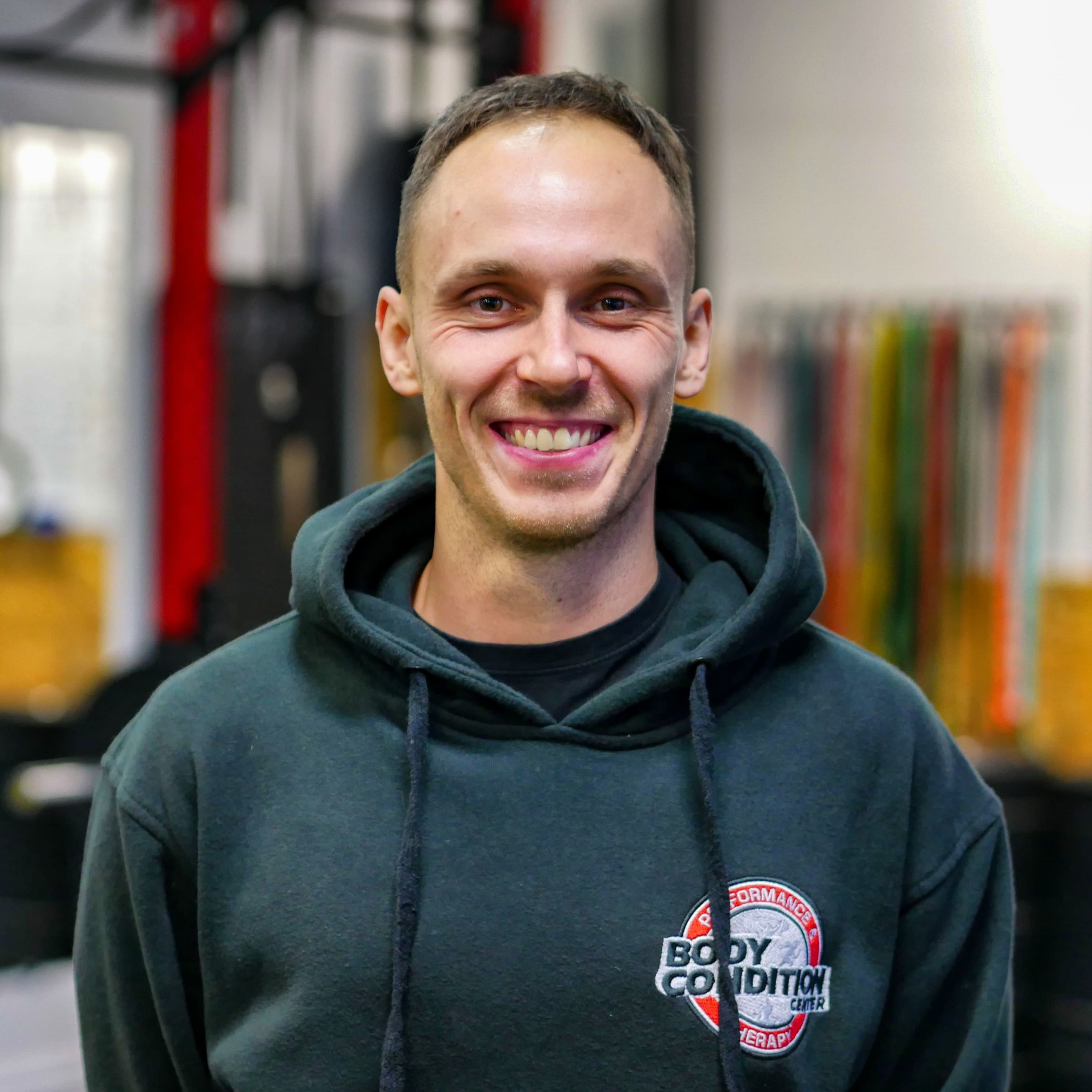 Uśmiechnięty mężczyzna w bluzie z kapturem z logo 'Body Condition Center' na tle sali treningowej z kolorowymi gumami do ćwiczeń.