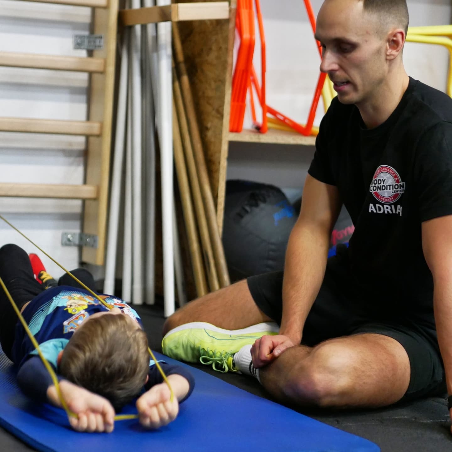 Chłopiec leżący na macie, ćwiczący z gumą pod nadzorem mężczyzny w czarnej koszulce z logo 'Body Condition Center', na tle drabinki gimnastycznej i sprzętu sportowego.