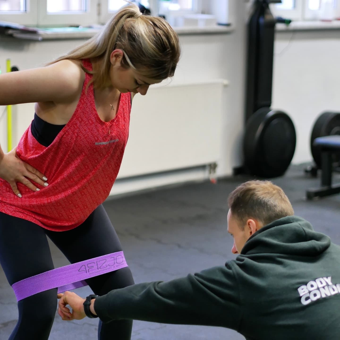 Kobieta w czerwonej koszulce ćwiczy z fioletową gumą na nogach pod okiem trenera w ciemnozielonej bluzie z napisem 'Body Condition' na plecach, w tle sprzęt do ćwiczeń.