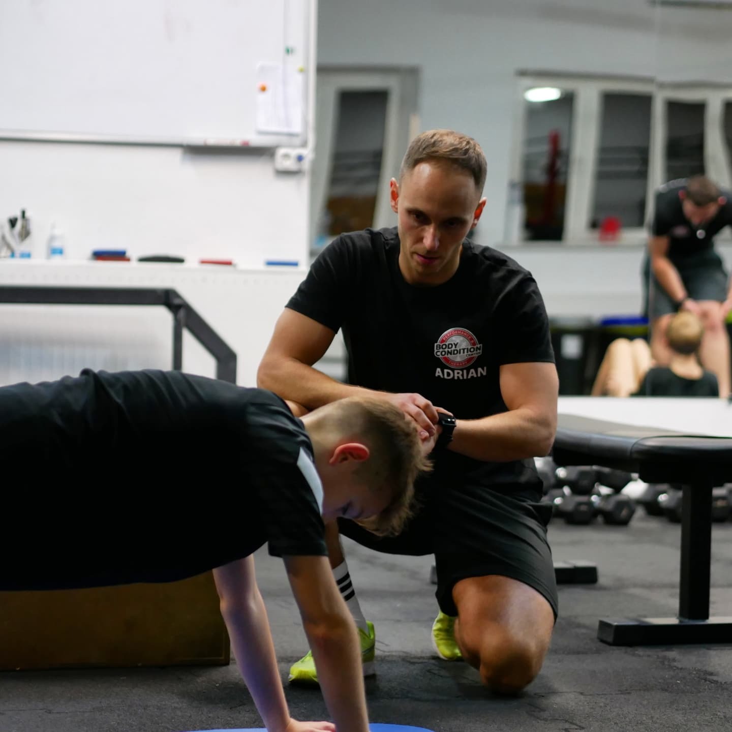Trener personalny Adrian nadzoruje chłopca wykonującego ćwiczenie plank na niebieskiej macie w siłowni; w tle inne osoby ćwiczą.